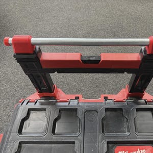 May include: A black and red Milwaukee tool box with a retractable silver handle. The handle is supported by red and black brackets. The tool box has several compartments and a red Milwaukee logo.