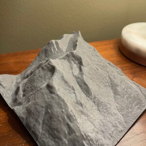 May include: A 3D printed model of a mountain range, with a textured surface and a gray color.