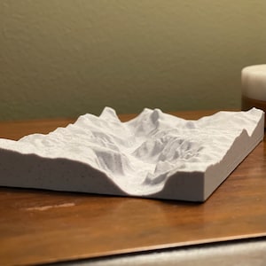 May include: A 3D-printed topographical model of a mountain range. The model is a light gray color and features detailed ridges, valleys, and peaks. The rectangular base sits on a wooden surface.