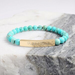 May include: A stretch bracelet featuring round turquoise-colored beads with dark veining and a central gold-toned metal bar. The bar is engraved with the cursive text Rooted & Rising. The beads are arranged in a continuous circular pattern.