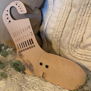 Könnte beinhalten: Eine Stricklehre aus Holz mit einem Haken zum Halten von Garn. Die Lehre hat Markierungen für verschiedene Nadelgrößen in US- und metrischen Größen. Die Lehre ist hellbraun und hat eine natürliche Holzoptik.