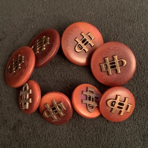 Peut inclure: Ensemble de neuf boutons ronds et ovales dans des tons de rouge et d'orange. Chaque bouton présente un emblème de couleur bronze. Les boutons sont disposés sur une surface sombre.