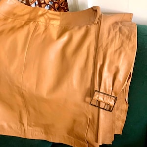 May include: A tan leather skirt with a matching belt and a rectangular buckle. The skirt is folded over, showing the texture and color of the leather. The skirt is resting on a dark green surface.