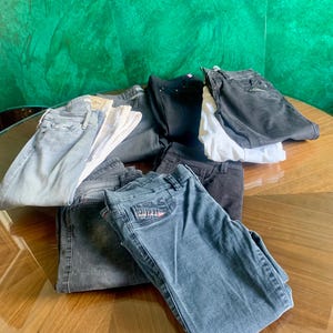 May include: A collection of folded denim jeans in various shades of blue, black, and gray. Some jeans have white fabric folded on top. The jeans are arranged on a wooden surface.
