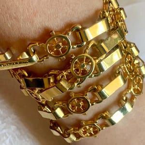 May include: A stack of gold-colored bracelets. Each bracelet features a series of rectangular links and small, circular charms with a wheel design. The bracelets are stacked on top of each other, creating a layered effect.
