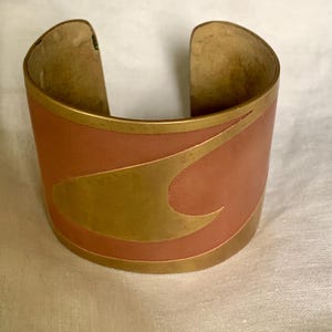 May include: A wide cuff bracelet with a copper-coloured base and a gold-coloured, curved design. The bracelet has gold trim along the top and bottom edges. The open design allows for easy wear.