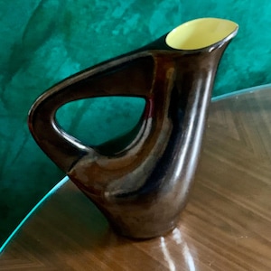 Puede incluir: Una jarra de cerámica marrón oscuro con un diseño de asa único. El interior de la jarra es de color amarillo claro. La jarra está sobre una superficie de madera con un fondo verde.