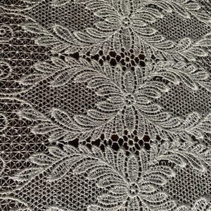 May include: Close-up of a white lace fabric with a floral pattern. The delicate design features repeating flower and leaf motifs, creating an elegant and textured surface. The lace is likely used for decorative purposes.