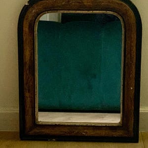 Puede incluir: Un espejo de pared arqueado con un marco de madera marrón oscuro y un borde interior marrón claro. El espejo tiene un cristal rectangular con las esquinas superiores redondeadas, que refleja una superficie verde azulada y un suelo claro.