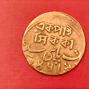 Op de afbeelding: Een gouden munt met Bengaalse tekst die "স্বর্ণ মুদ্রা" luidt en Arabische tekst. De munt ligt op een rood oppervlak.