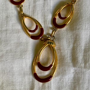 Peut inclure: Collier doré avec des maillons ovales et un accent en émail rouge foncé. Le pendentif présente un double ovale avec un émail rouge assorti. Le bijou est présenté sur une surface blanche texturée.