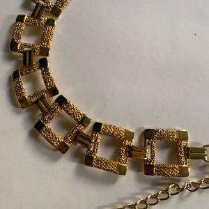 Puede incluir: Un cinturón dorado con una serie de eslabones cuadrados texturizados. El cinturón tiene una extensión de cadena para ajustar la talla. Los eslabones tienen una superficie texturizada, y el diseño general es elegante y clásico.