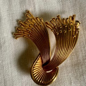 Peut inclure: Broche dorée au design abstrait unique. La broche présente une forme tourbillonnante en forme de ruban avec des détails texturés et striés et de petits accents arrondis. L'esthétique générale est élégante et d'inspiration vintage.