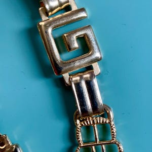 Puede incluir: Primer plano de un cinturón de metal dorado con un diseño de llave griega. El cinturón presenta eslabones rectangulares y cuadrados, con un patrón geométrico texturizado. El fondo es de color azul claro sólido.