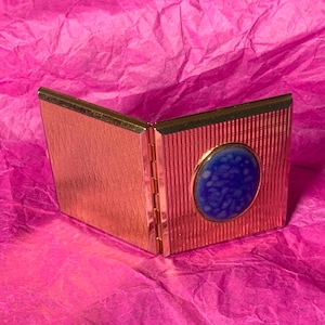May include: A gold-toned, hinged compact with a textured finish. The compact features a blue oval-shaped embellishment with a speckled design. The open compact is displayed on a pink, textured surface.
