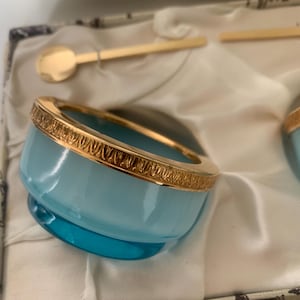 May include: A light blue glass bowl with a gold-toned metal rim featuring a decorative leaf pattern. The bowl is accompanied by a small gold-toned metal spoon, both presented within a fabric-lined container with a patterned exterior.
