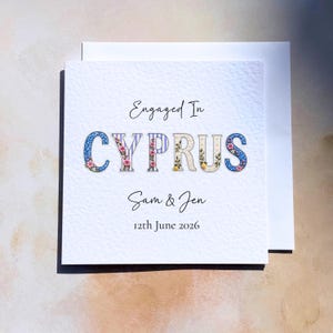 May include: A square white greeting card featuring the text Engaged In CYPRUS in a decorative floral and striped font. Below the word CYPRUS, the names Sam & Jen are printed in a script font, followed by the date 12th June 2026 in a simple font.