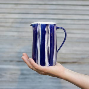 Könnte beinhalten: Ein blauer und weiß gestreifter Keramik Krug mit Henkel. Der Krug ist handbemalt mit weißen Streifen auf einem dunkelblauen Hintergrund.