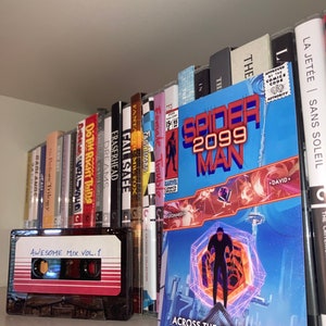 Pu&ograve; includere: Una mensola mostra una musicassetta con l'etichetta "AWESOME MIX VOL. 1" accanto a un fumetto di "Spider-Man 2099". Una collezione di film Criterion Collection e altri libri sono visibili sullo sfondo.