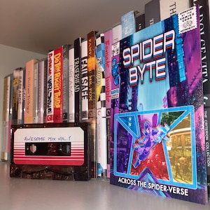 Pode incluir: Uma estante com uma variedade de livros e uma fita cassete com a etiqueta "Awesome Mix Vol. 1". Um gibi intitulado "Spider Byte: Across the Spider-Verse" é exibido de forma proeminente na estante.