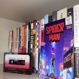 Puede incluir: Una estanter&iacute;a llena de libros variados y una cinta de casete etiquetada como "Awesome Mix Vol. 1". La estanter&iacute;a est&aacute; llena de libros sobre diversos temas, incluyendo cine, literatura y c&oacute;mics. El libro m&aacute;s destacado es un c&oacute;mic de Spider-Man con el t&iacute;tulo "Across the Spider-Verse".