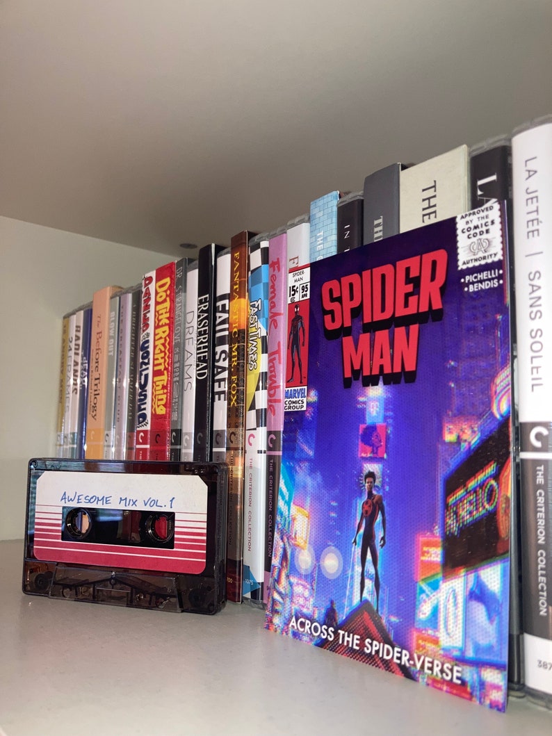 Puede incluir: Una estanter&iacute;a llena de libros variados y una cinta de casete etiquetada como "Awesome Mix Vol. 1". La estanter&iacute;a est&aacute; llena de libros sobre diversos temas, incluyendo cine, literatura y c&oacute;mics. El libro m&aacute;s destacado es un c&oacute;mic de Spider-Man con el t&iacute;tulo "Across the Spider-Verse".