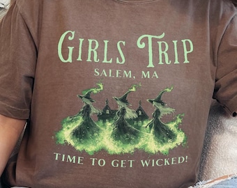 May include: A brown t-shirt with a green graphic of three witches flying over houses with the text "Girls Trip Salem, MA Time to Get Wicked!"