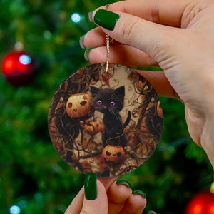 Può includere: Ornamento natalizio rotondo in ceramica con un gatto nero con occhi viola e due lanterne di zucca. L'ornamento ha un cordino dorato per appenderlo. Lo sfondo è un'immagine sfocata di un albero di Natale.