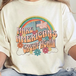 May include: A white t-shirt with a retro-style graphic featuring a rainbow, an American flag, and the text "Make Americans Great Again".