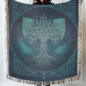 May include: A woven throw blanket with a dark blue background and a green tree design. The tree is surrounded by a circle and has a stylized axe design at the top. The blanket has a fringe border.