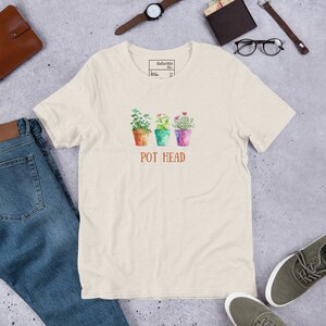 Puede incluir: Una camiseta beige claro con un gráfico de tres plantas en maceta con flores de colores y el texto "POT HEAD" en letras marrones.