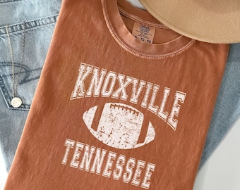 May include: A rust-colored t-shirt with a distressed white graphic of a football and the text "Knoxville Tennessee".