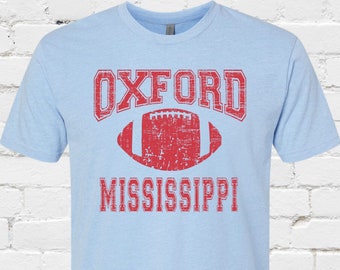 May include: Pale blue t-shirt with a distressed red design. The design includes the word "OXFORD" above a football graphic and the word "MISSISSIPPI" below. The shirt is a heather columbia blue colour and made of a soft material.