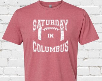 May include: A red heather t-shirt with a white graphic that says "Saturday in Columbus" with a football graphic inside the text.