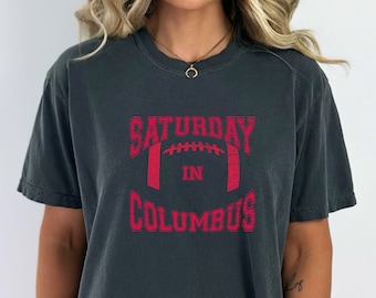 May include: A dark gray t-shirt with a red graphic that says "Saturday in Columbus" with a football graphic in the middle.