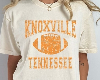 May include: A white t-shirt with an orange distressed graphic of a football and the text "KNOXVILLE TENNESSEE". The shirt is shown on a model wearing black biker shorts. The color of the shirt is ivory.