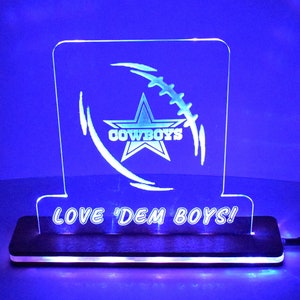 May include: A clear acrylic sign with a Dallas Cowboys logo and the words "LOVE 'DEM BOYS!"  The sign is illuminated with blue LED lights and sits on a wooden base.