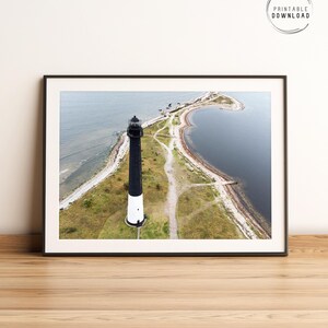 Può includere: Vista aerea di un faro bianco e nero su una piccola isola. Il faro è circondato da erba verde e un'area d'acqua.