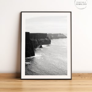 Może przedstawiać: Czarno-białe zdjęcie dramatycznego klifu górującego nad rozległym oceanem. Klify są wysokie i skaliste, z nutą mgły w powietrzu. Ocean jest spokojny i majestatyczny, z kilkoma falami rozbijającymi się o brzeg.