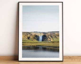May include: A photograph of a waterfall cascading down a green hillside, with a still body of water in the foreground. The sky is a light blue with white clouds.