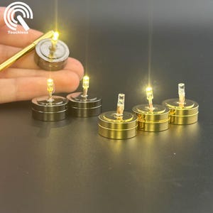 Op de afbeelding: Een verzameling kleine, batterijgevoede LED-lampjes met een warme, gele gloed. De lampjes zijn cilindrisch, met een metalen basis en een kleine, heldere LED-lamp. Sommige zijn zilverkleurig, andere goudkleurig. Een goudkleurige staaf is ook aanwezig.