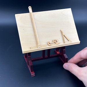Könnte beinhalten: Ein kleiner Holz-Zeichentisch mit rotem Untergestell und einer hellbraunen Tischplatte. Der Tisch enthält ein Lineal, einen Winkelmesser und ein gebogenes Zeichenwerkzeug.