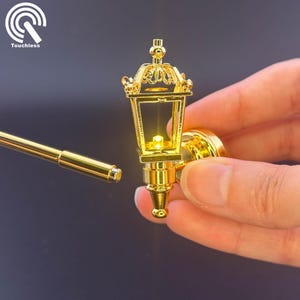 Op de afbeelding: Een miniatuur gouden wandlamp met een lichtje erin. De lamp heeft een decoratief ontwerp en is verlicht.