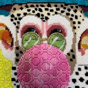 May include: A close-up of a needlepoint design of a monkey wearing sunglasses and blowing a pink bubble. The monkey has a black and white spotted fur pattern and is wearing a blue and red hat.