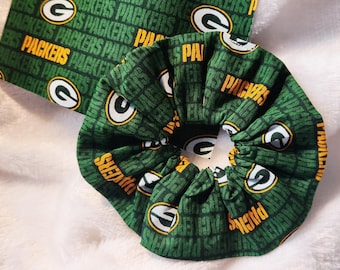 Puede incluir: Coletero verde y dorado con el texto "PACKERS" y un logotipo "G" repetido en la tela. Se ve un trozo de tela a juego. El coletero es un accesorio para el pelo.