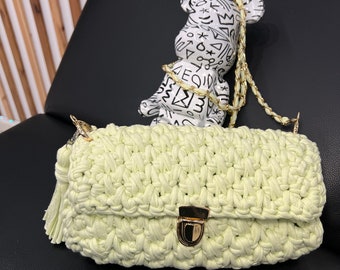 Könnte beinhalten: Eine hellgrüne gehäkelte Handtasche mit einem goldenen Verschluss und einem Quastenanhänger. Die Tasche hat einen Kettenriemen und liegt auf einer schwarzen Oberfläche. Ein weißer Keramikbär mit schwarzen Gekritzel ist auf der Tasche.