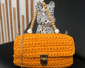 Könnte beinhalten: Eine leuchtend orangefarbene gehäkelte Handtasche mit einer goldenen Kettenkette und einem goldenen Verschluss. Die Tasche liegt auf einer schwarzen Lederoberfläche.