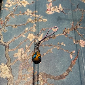 May include: A small, round, orange and blue charm with a silver loop attached to a pink string. The charm is on a blue background with a floral pattern.