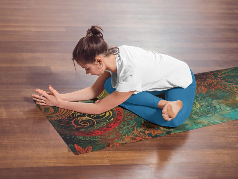 May include: A woman sits on a colorful yoga mat with intricate patterns in shades of green, red, yellow, and blue. She is wearing blue yoga pants and a white t-shirt.
