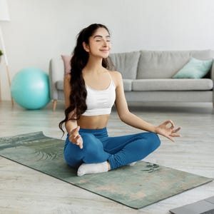 May include: A woman sits in a yoga pose on a blue and green patterned yoga mat. She is wearing a white sports bra and blue yoga pants. She has long dark hair and is looking up with her eyes closed.